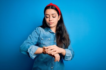 Young brunette woman wearing casual denim shirt over blue isolated background Checking the time on wrist watch, relaxed and confident
