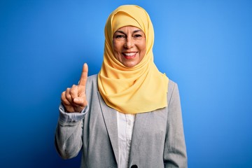 Middle age brunette business woman wearing muslim traditional hijab over blue background pointing finger up with successful idea. Exited and happy. Number one.