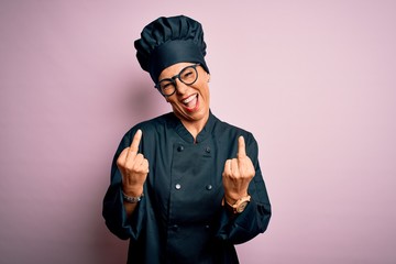 Middle age brunette chef woman wearing cooker uniform and hat over isolated pink background Showing middle finger doing fuck you bad expression, provocation and rude attitude. Screaming excited