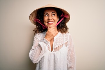 Middle age brunette woman wearing asian traditional conical hat over white background with hand on chin thinking about question, pensive expression. Smiling with thoughtful face. Doubt concept.