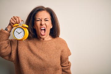 Middle age brunette woman holding clasic alarm clock over isolated background angry and mad...