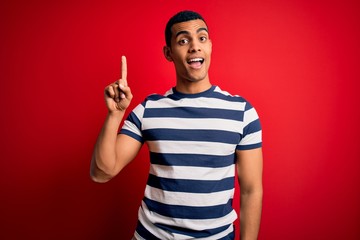Naklejka premium Handsome african american man wearing casual striped t-shirt standing over red background pointing finger up with successful idea. Exited and happy. Number one.