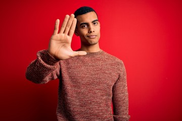 Young handsome african american man wearing casual sweater standing over red background doing stop sing with palm of the hand. Warning expression with negative and serious gesture on the face.