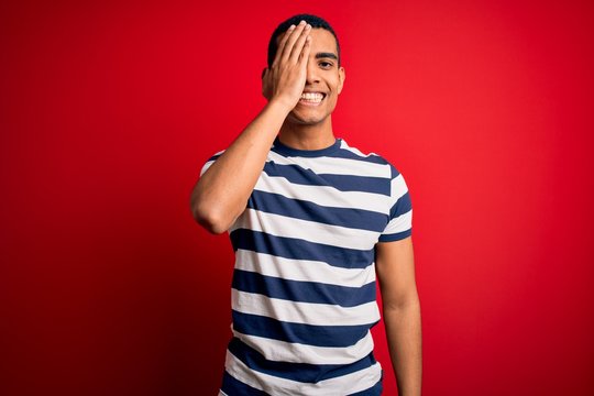 Handsome african american man wearing casual striped t-shirt standing over red background covering one eye with hand, confident smile on face and surprise emotion.