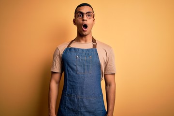 Young handsome african american shopkeeper man wearing apron over yellow background afraid and...