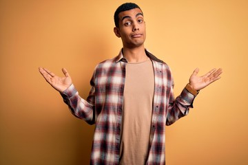 Young handsome african american man wearing casual shirt standing over yellow background clueless and confused expression with arms and hands raised. Doubt concept.