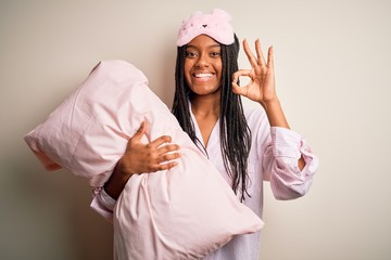 Young african american woman wearing pajama and eye mask hugging bed pillow doing ok sign with fingers, excellent symbol