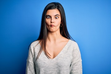Young beautiful brunette woman wearing casual sweater standing over blue background making fish face with lips, crazy and comical gesture. Funny expression.