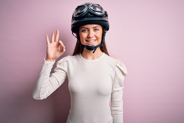 Young beautiful motorcyclist woman with blue eyes wearing moto helmet over pink background smiling positive doing ok sign with hand and fingers. Successful expression.