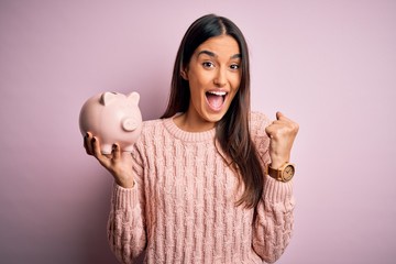 Young beautiful brunette woman holding piggy bank saving money for retirement screaming proud and...