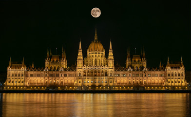 Fototapeta na wymiar Budapest Parliament Building illuminated at night with dark sky, giant full moon, reflection in Danube river and river with silk texture.