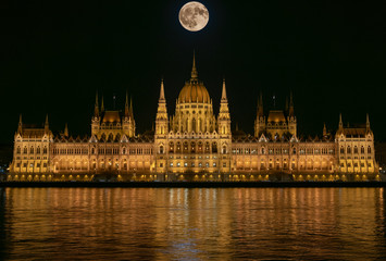 Fototapeta premium Budapest Parliament Building illuminated at night with dark sky, giant full moon, reflection in Danube river and river with silk texture.