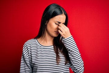 Young beautiful brunette woman wearing casual striped t-shirt over red background tired rubbing nose and eyes feeling fatigue and headache. Stress and frustration concept.