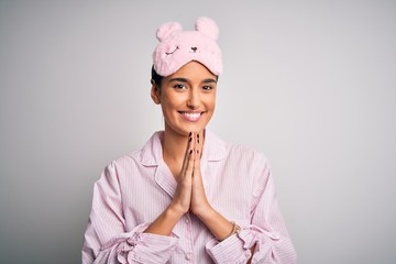 Young beautiful brunette woman wearing pajama and sleep mask over white background praying with hands together asking for forgiveness smiling confident.