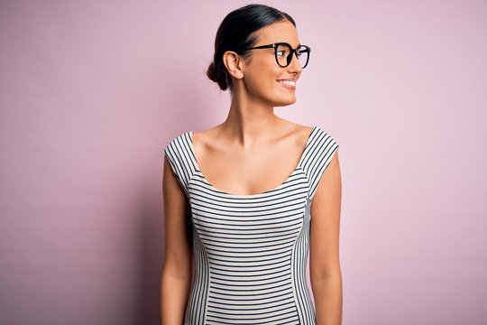 Young beautiful woman wearing casual striped dress and glasses over pink background looking away to side with smile on face, natural expression. Laughing confident.