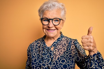 Senior beautiful grey-haired woman wearing casual shirt and glasses over yellow background doing happy thumbs up gesture with hand. Approving expression looking at the camera showing success. - obrazy, fototapety, plakaty