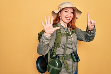 Young redhead backpacker woman hiking wearing backpack and hat over yellow background showing and...