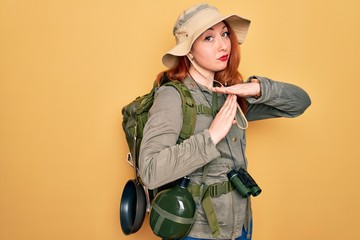 Young redhead backpacker woman hiking wearing backpack and hat over yellow background Doing time...