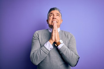 Middle age handsome grey-haired man wearing elegant sweater over purple background begging and praying with hands together with hope expression on face very emotional and worried. Begging.