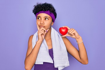 Beautiful african american afro sporty woman doing exercise wearing towel holding heart serious...