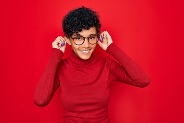 Fototapeta na wymiar Young beautiful african american afro woman wearing turtleneck sweater and glasses Smiling pulling ears with fingers, funny gesture. Audition problem