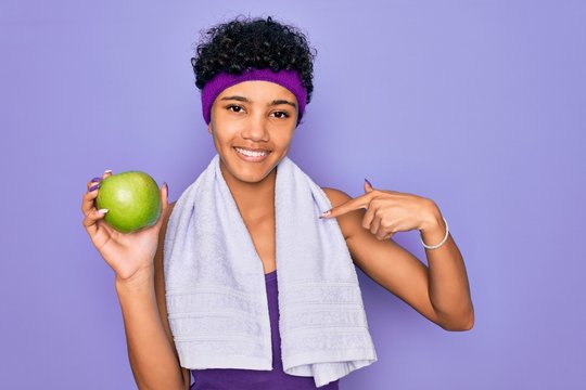 Beautiful african american afro sporty woman doing exercise wearing towel eating green apple with surprise face pointing finger to himself
