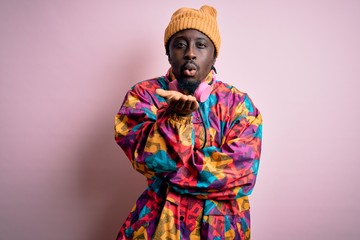 Young handsome african american man wearing colorful coat and cap over pink background looking at the camera blowing a kiss with hand on air being lovely and sexy. Love expression.