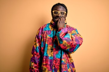 Handsome african american man wearing colorful coat and glasses over yellow background looking stressed and nervous with hands on mouth biting nails. Anxiety problem.