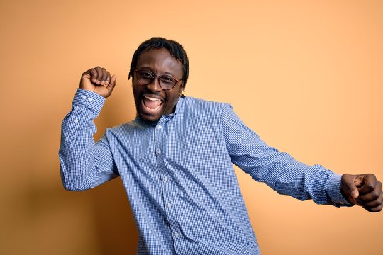 Young handsome african american man wearing shirt and glasses over yellow background Dancing happy and cheerful, smiling moving casual and confident listening to music