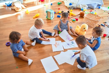 Adorable group of toddlers sitting on the floor drawing using paper and pencil around lots of toys...