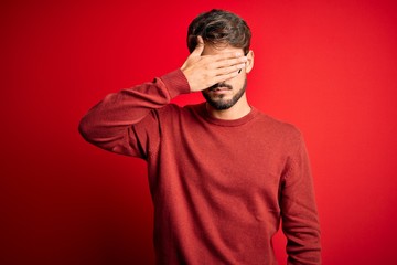 Young handsome man with beard wearing glasses and sweater standing over red background covering eyes with hand, looking serious and sad. Sightless, hiding and rejection concept