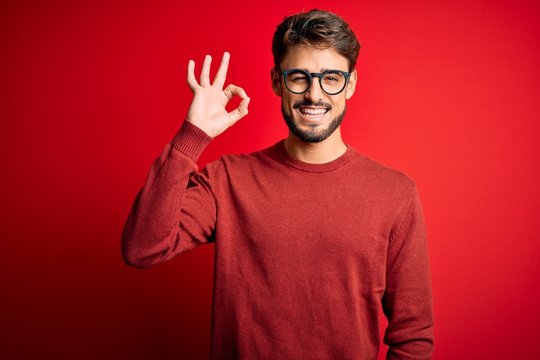 Young handsome man with beard wearing glasses and sweater standing over red background smiling positive doing ok sign with hand and fingers. Successful expression.