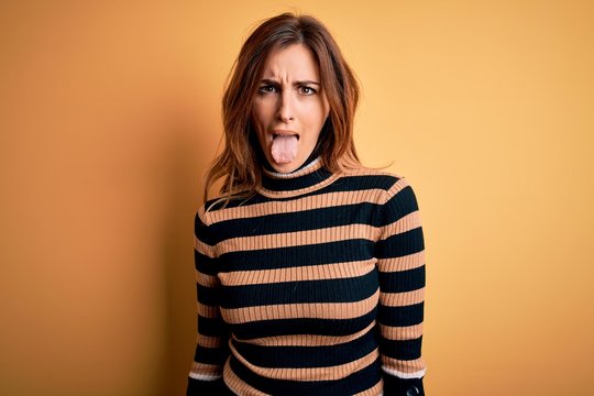 Young beautiful brunette woman wearing striped turtleneck sweater over yellow background sticking tongue out happy with funny expression. Emotion concept.
