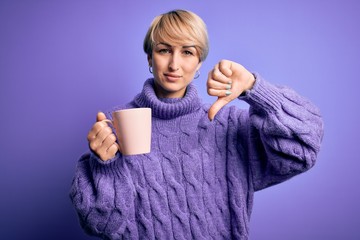 Naklejka premium Young blonde woman with short hair wearing winter sweater drinking a cup of hot coffee with angry face, negative sign showing dislike with thumbs down, rejection concept