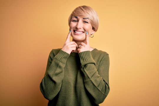 Young blonde woman with modern short hair wearing casual sweater over yellow background Smiling with open mouth, fingers pointing and forcing cheerful smile