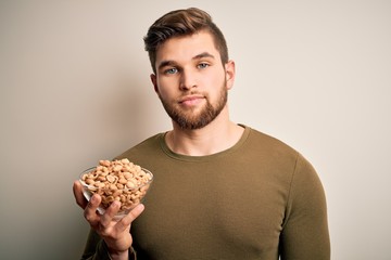 Young blond man with beard and blue eyes holding bowl with healthy salty peanuts with a confident expression on smart face thinking serious