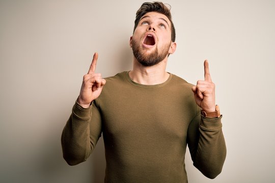Young blond man with beard and blue eyes wearing green sweater over white background amazed and surprised looking up and pointing with fingers and raised arms.