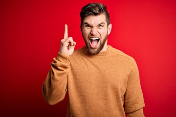 Young blond man with beard and blue eyes wearing casual sweater over red background pointing finger up with successful idea. Exited and happy. Number one.