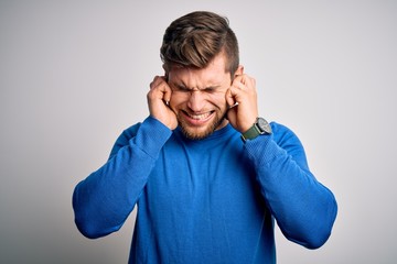 Young handsome blond man with beard and blue eyes wearing casual sweater covering ears with fingers with annoyed expression for the noise of loud music. Deaf concept.