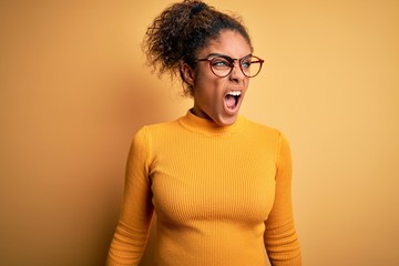 Young beautiful african american girl wearing sweater and glasses over yellow background angry and mad screaming frustrated and furious, shouting with anger. Rage and aggressive concept.