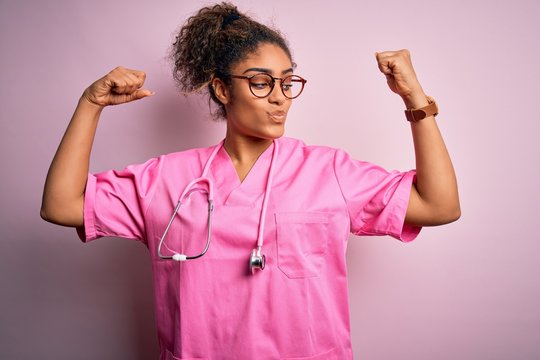African american nurse girl wearing medical uniform and stethoscope over pink background showing arms muscles smiling proud. Fitness concept.