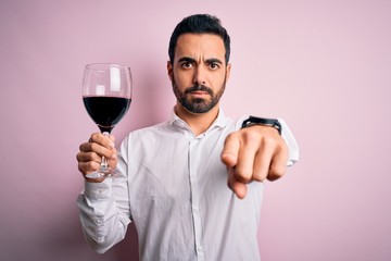 Young handsome man with beard drinking glass of red wine over isolated pink background pointing with finger to the camera and to you, hand sign, positive and confident gesture from the front