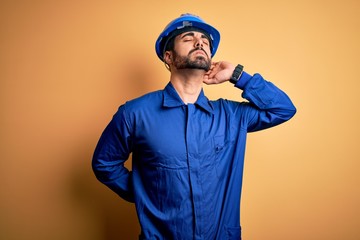 Mechanic man with beard wearing blue uniform and safety helmet over yellow background Suffering of neck ache injury, touching neck with hand, muscular pain