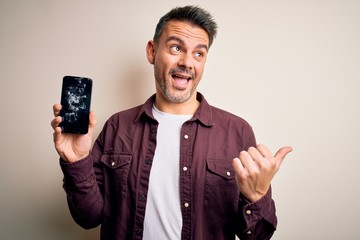 Young handsome man holding broken smartphone showing craked screen pointing and showing with thumb...