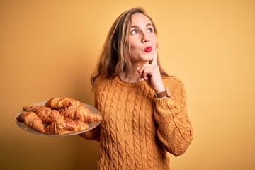 Young beautiful blonde woman holding plate with croissants over isolated yellow background serious...