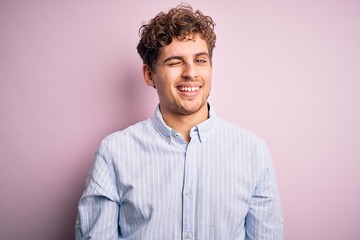 Young blond handsome man with curly hair wearing striped shirt over white background winking...