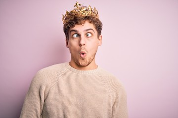 Young blond man with curly hair wearing golden crown of king over pink background making fish face...