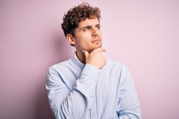 Young blond handsome man with curly hair wearing striped shirt over white background with hand on chin thinking about question, pensive expression. Smiling with thoughtful face. Doubt concept.