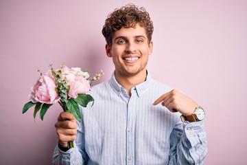 Young blond man with curly hair holding beautiful bouquet over isolated pink background with surprise face pointing finger to himself