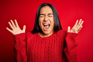 Young beautiful asian woman wearing casual sweater standing over isolated red background celebrating mad and crazy for success with arms raised and closed eyes screaming excited. Winner concept